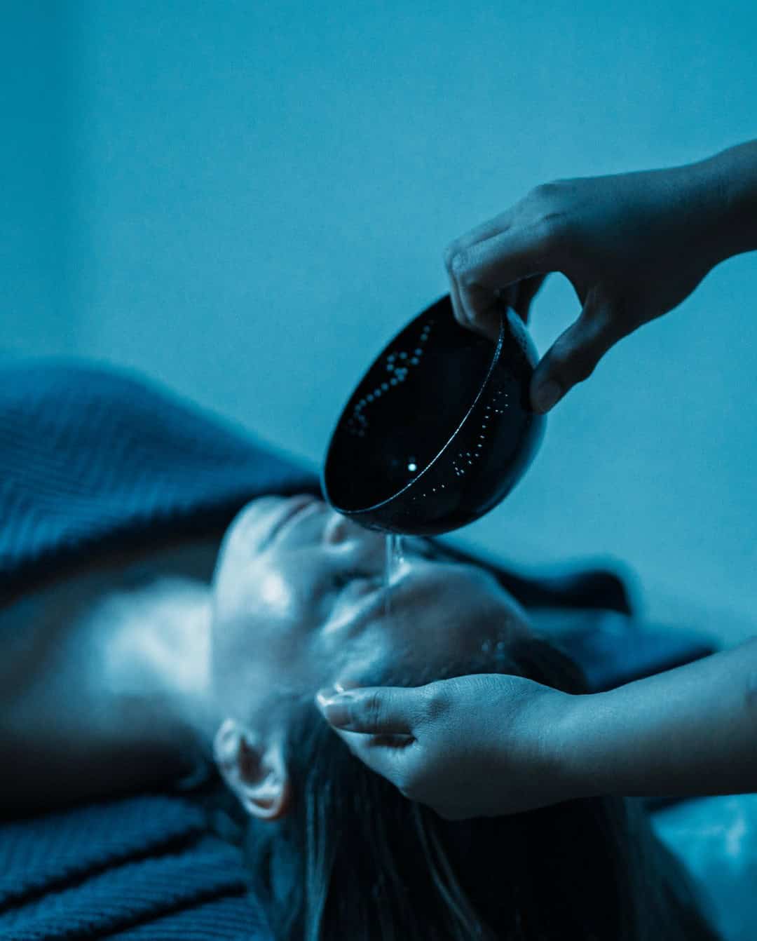 Person lying down with water being gently poured onto their forehead from a black bowl in a tranquil spa setting, embodying the essence of Grand Cayman Luxury Travel.