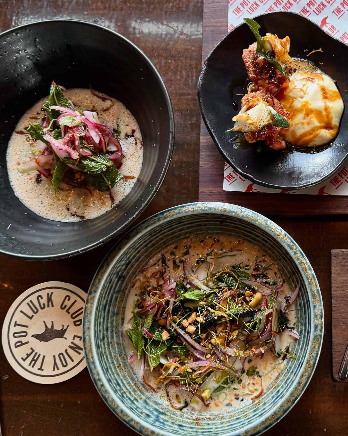 Three bowls of colorful, garnished dishes with fresh herbs sit on a wooden table next to a coaster, perfect for exploring new foodie destinations.
