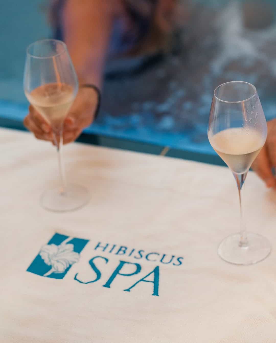 Two glasses of champagne rest on a Hibiscus Spa towel near a sparkling pool, with a person reaching for a glass&mdash;an inviting scene that perfectly captures the essence of Grand Cayman Luxury Travel.