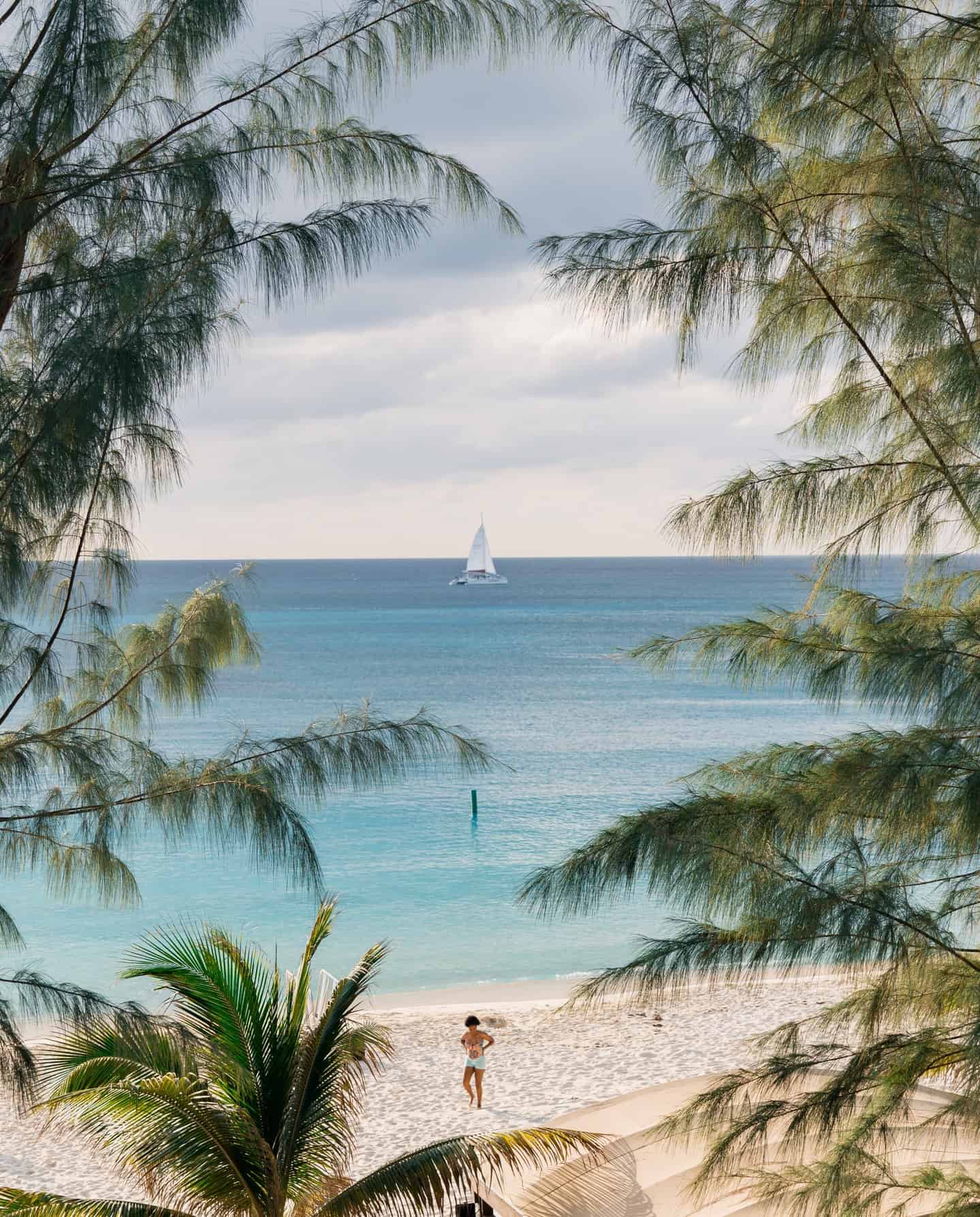 A sailboat on the ocean, viewed through trees, with a person standing on a sandy beach in the foreground&mdash;a scene that captures the essence of Grand Cayman luxury travel.