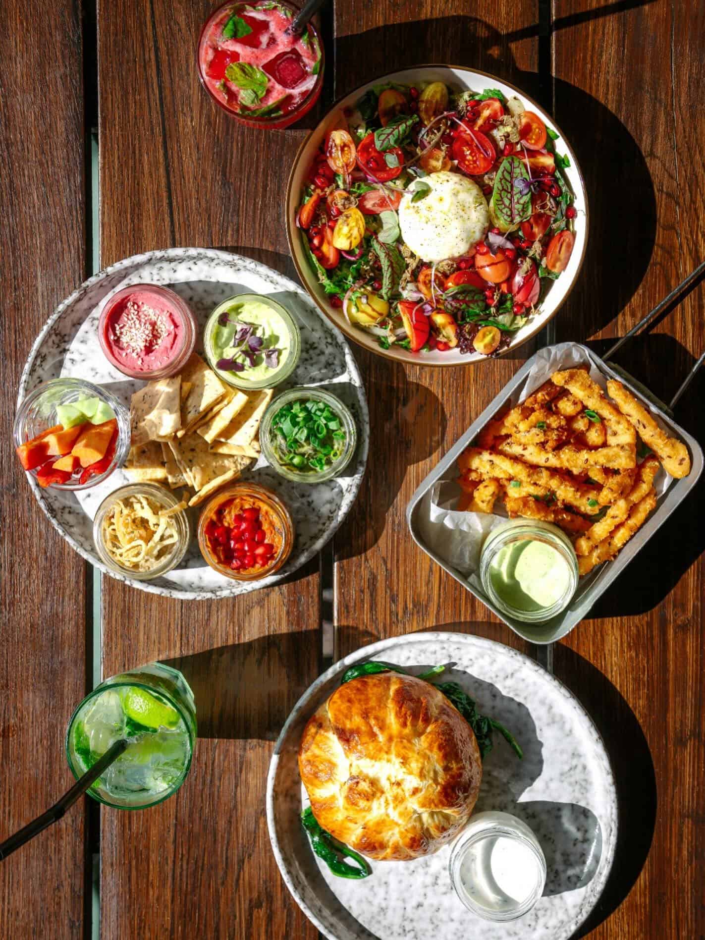 Colorful bowls of salad, dips, fries, drinks, and a pastry dish on a wooden table in sunlight—perfect for exploring top Foodie Destinations.