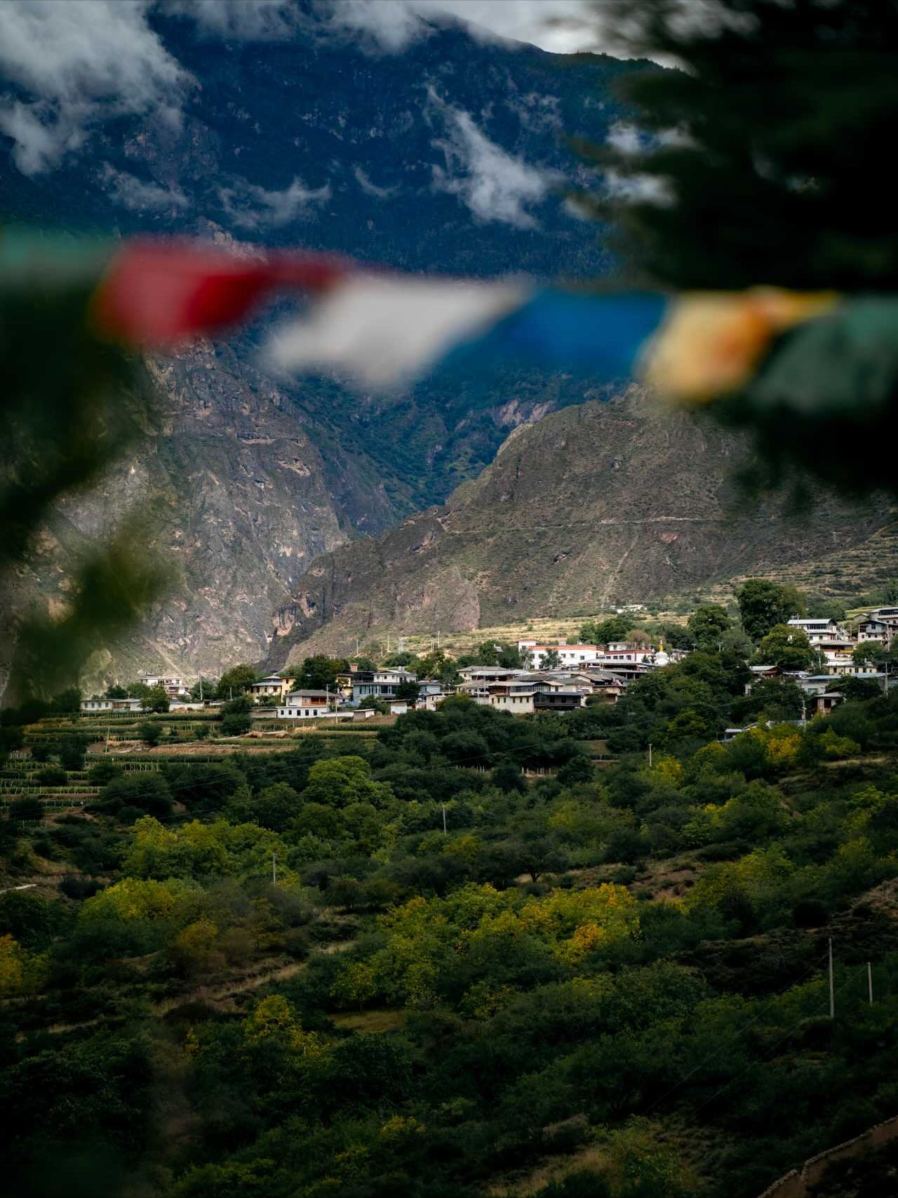 Colorful prayer flags blurred in the foreground, with a mountain village known for wine tourism nestled among lush green hills in the background.