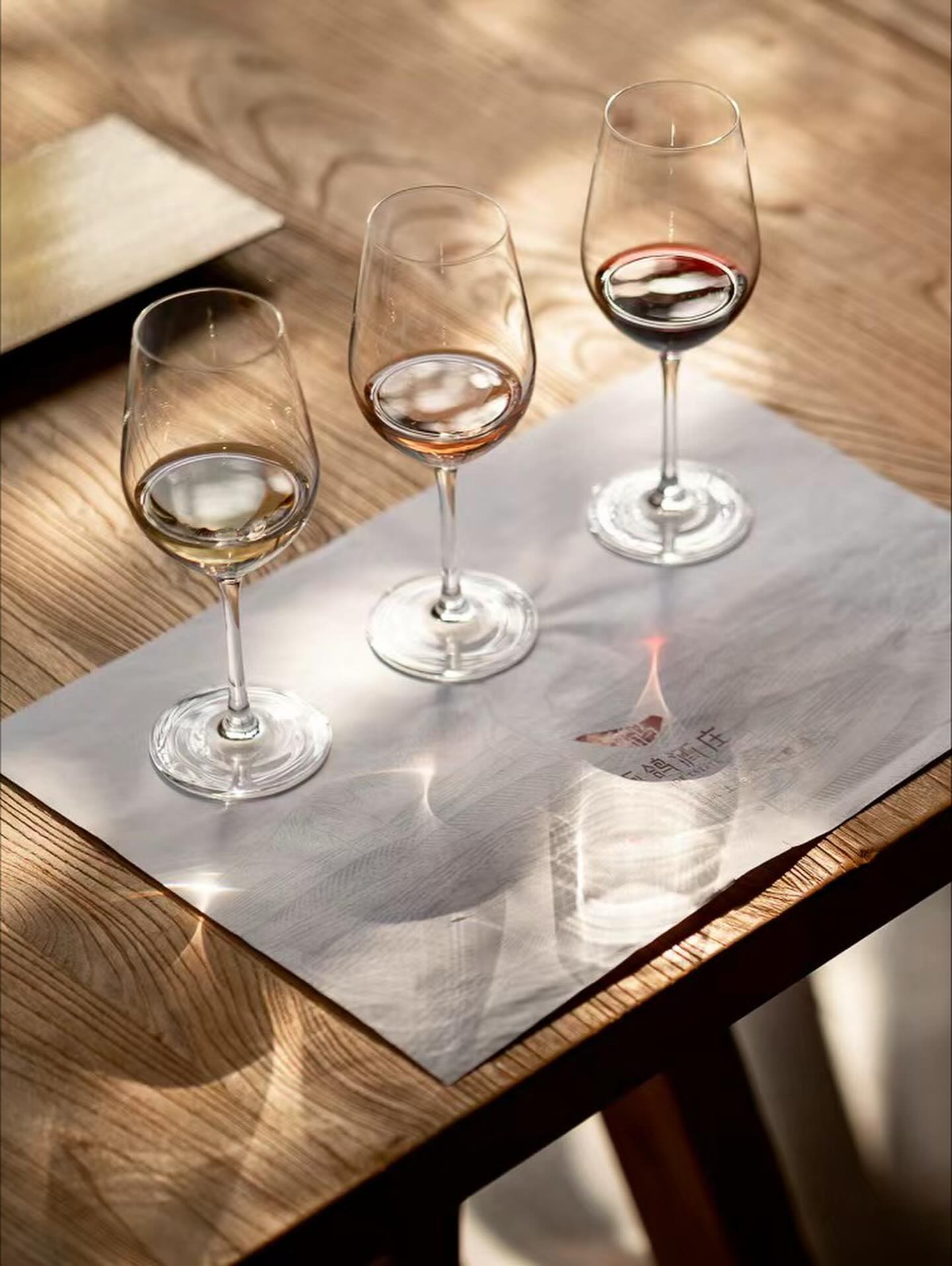 Three wine glasses with different amounts of wine on a light placemat rest on a wooden table in sunlight, evoking the essence of wine tourism.