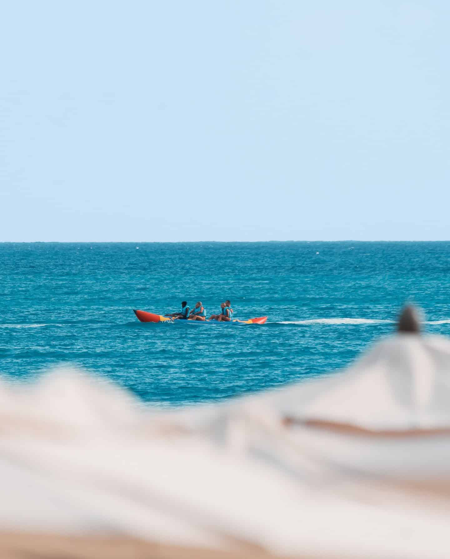 Three people paddle a red kayak on blue ocean water, with blurred beach umbrellas in the foreground&mdash;an idyllic moment that captures the allure of Grand Cayman luxury travel.