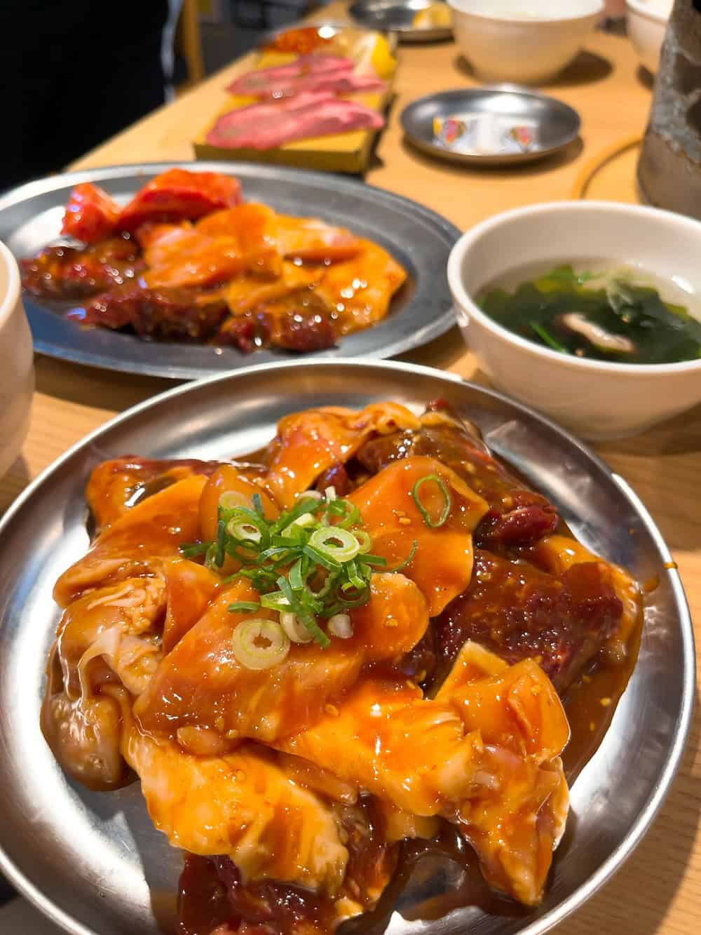 Plates of marinated raw meat garnished with green onions, set against bowls of soup and more dishes in the background—perfect for those seeking top foodie destinations.
