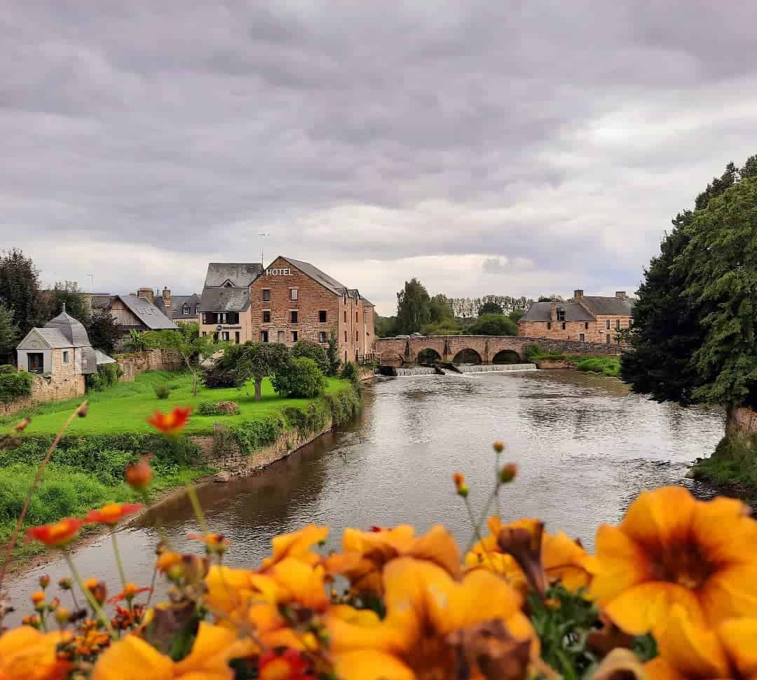 A stone hotel by a river, with an arched bridge and orange flowers in the foreground under a cloudy sky—perfect scenery for a luxury barge cruise nearby.