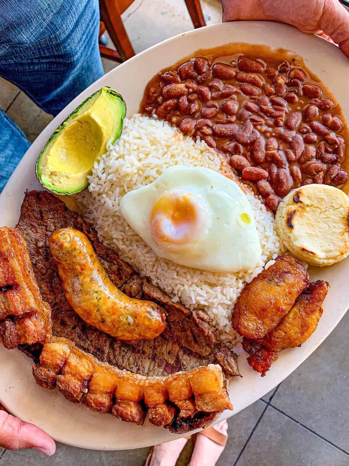 A plate perfect for Foodie Destinations, featuring rice, beans, fried egg, steak, sausage, avocado, plantains, arepa, and pork belly.