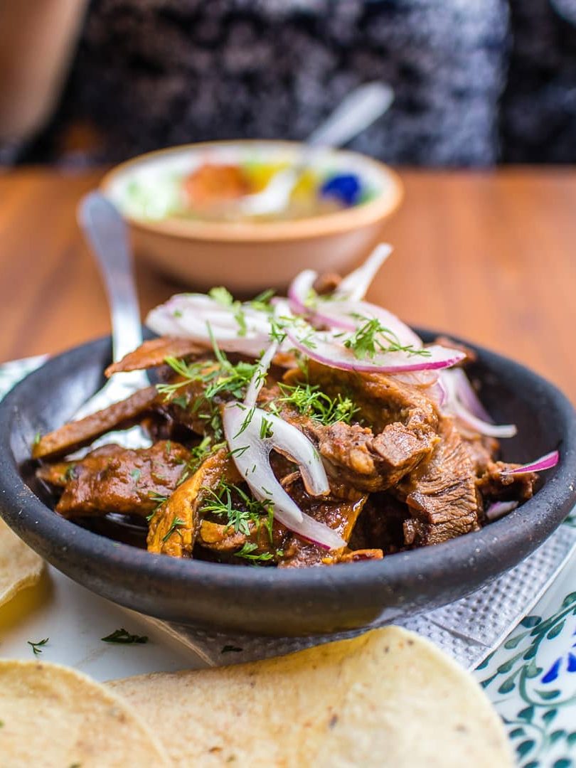 A bowl of beef strips with onions and herbs, served with tortillas on a patterned plate—perfect for those seeking authentic flavors and foodie destinations.
