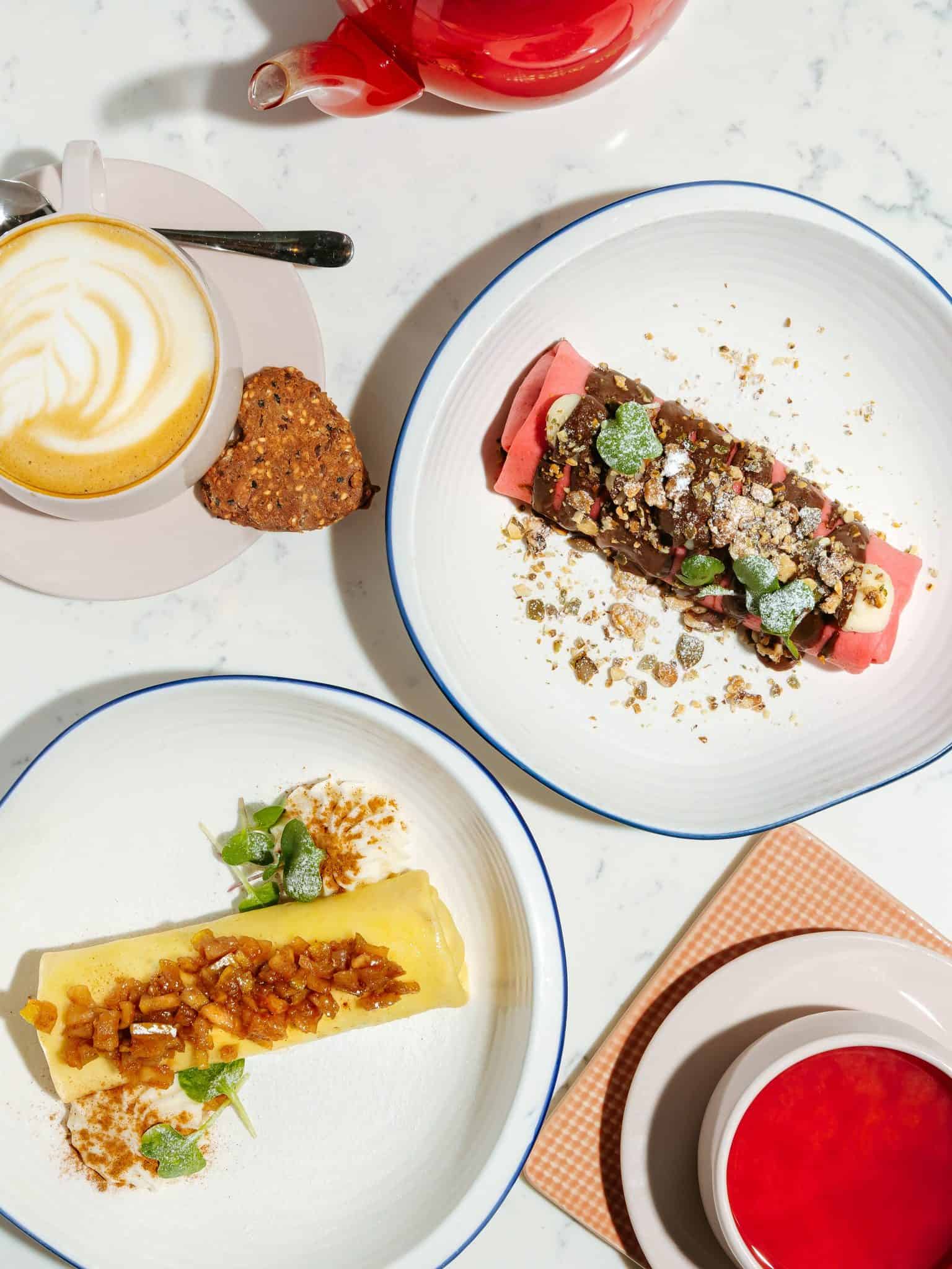 Two plates of rolled crepes, a cappuccino with biscotti, and a cup of bright red tea on a white table make this spot one of the must-visit foodie destinations.