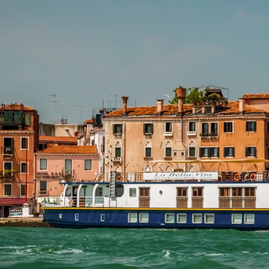 A white and blue boat named "La Bella Vita," offering a luxury barge cruise, is docked by colorful buildings along a canal.