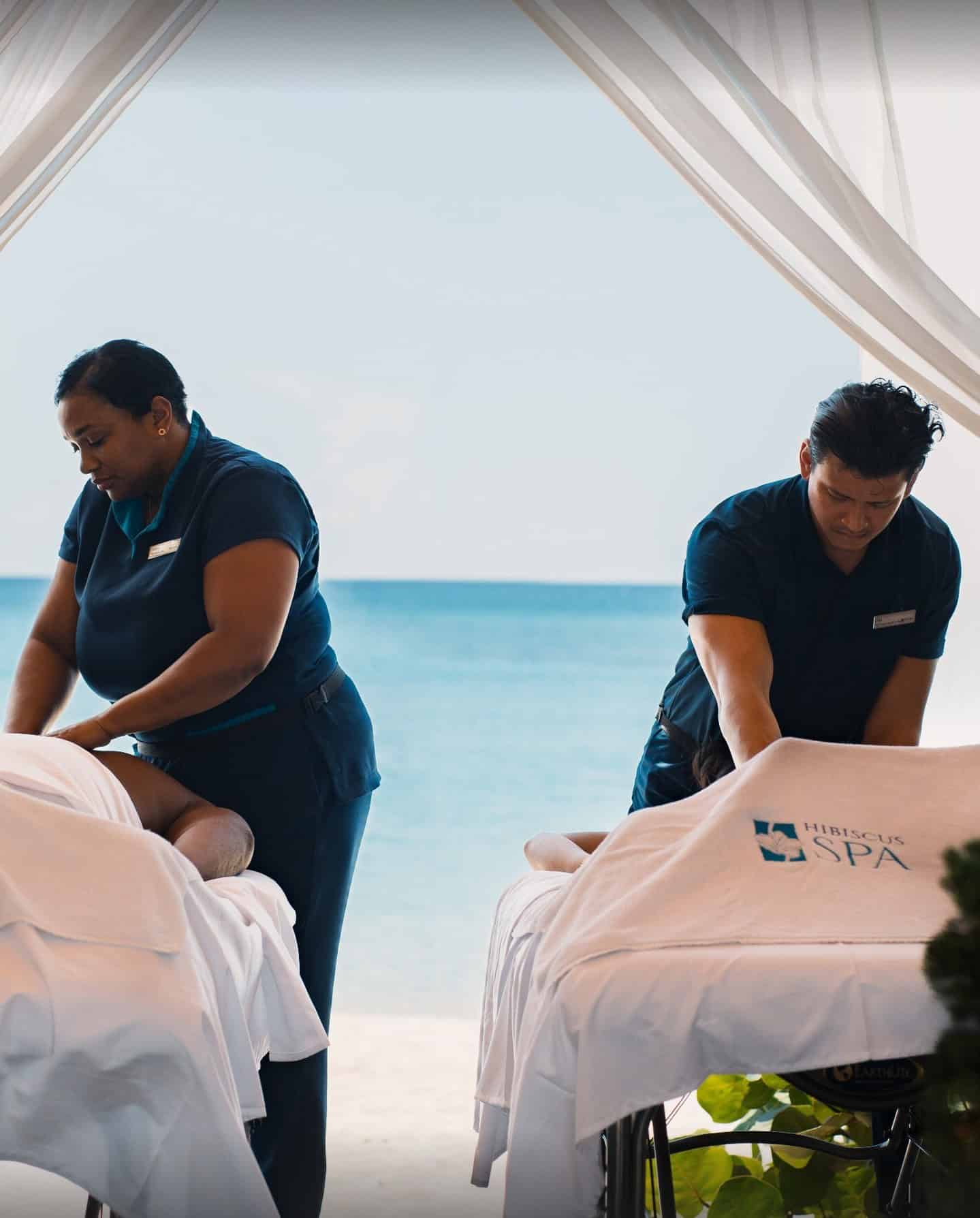 Two massage therapists give massages on tables by the ocean under white drapes, with a spa logo visible, offering the ultimate Grand Cayman Luxury Travel experience.