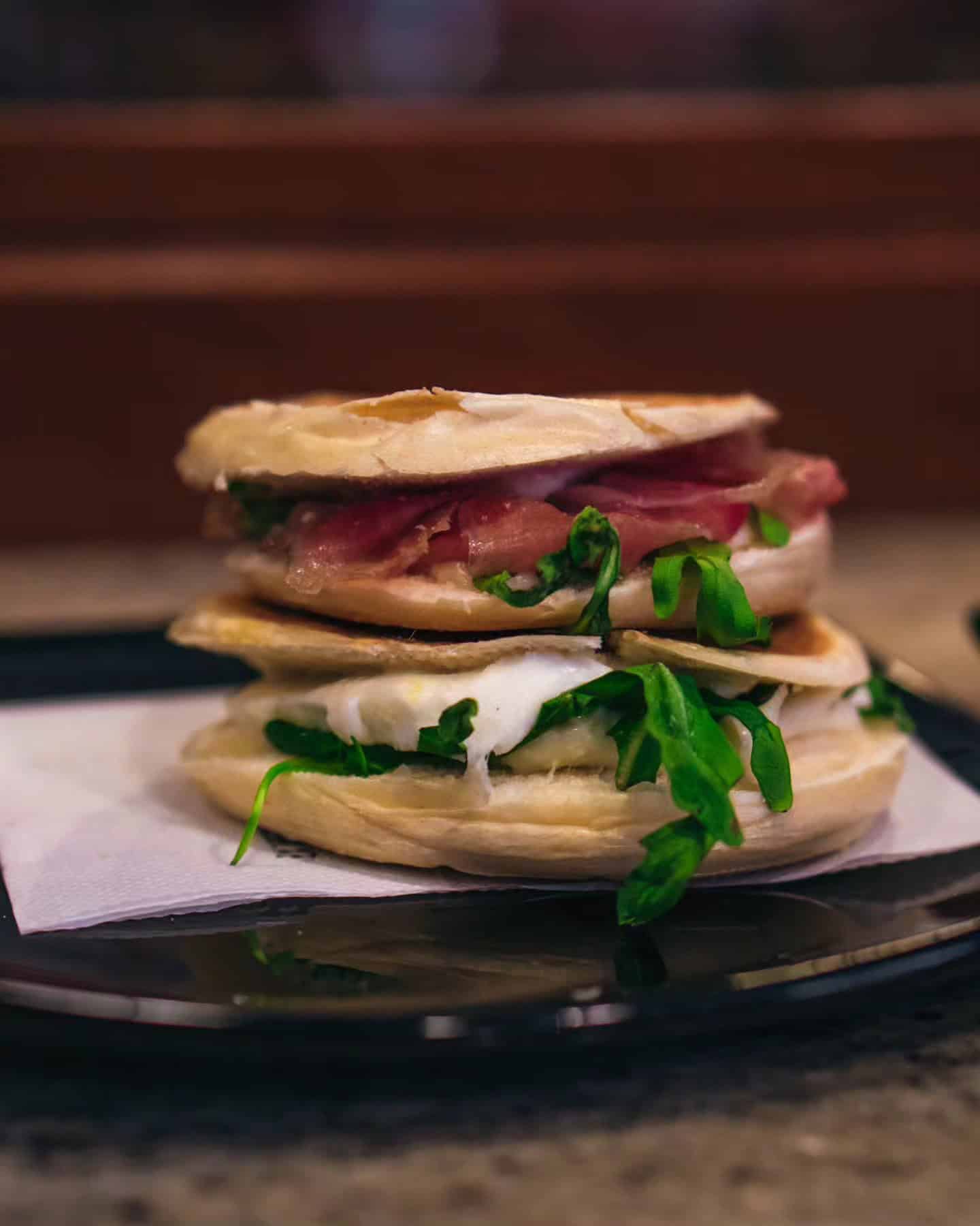Two stacked sandwiches with greens, cheese, and meat sit on a black plate with a napkin underneath—perfect for those seeking new foodie destinations.