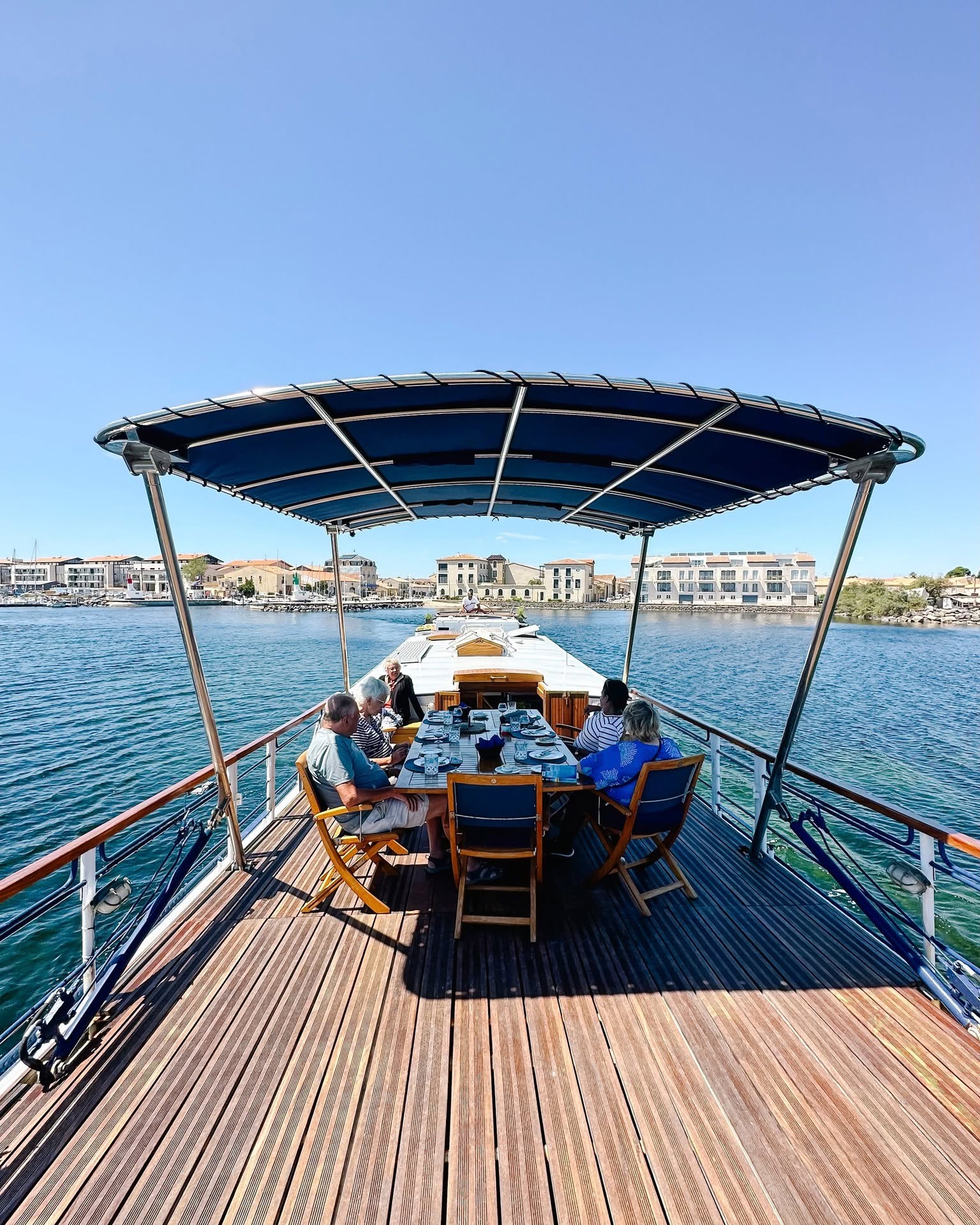 Four people sit at a set table on a boat deck under a canopy, enjoying a luxury barge cruise with water and buildings in the background.