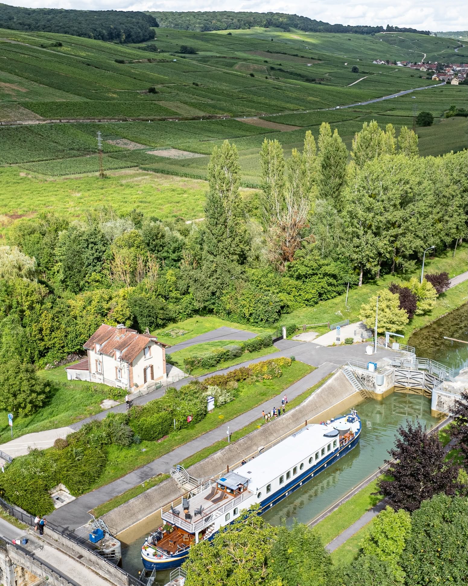 A luxury barge cruise passes through a lock near a small house, surrounded by green fields and trees.