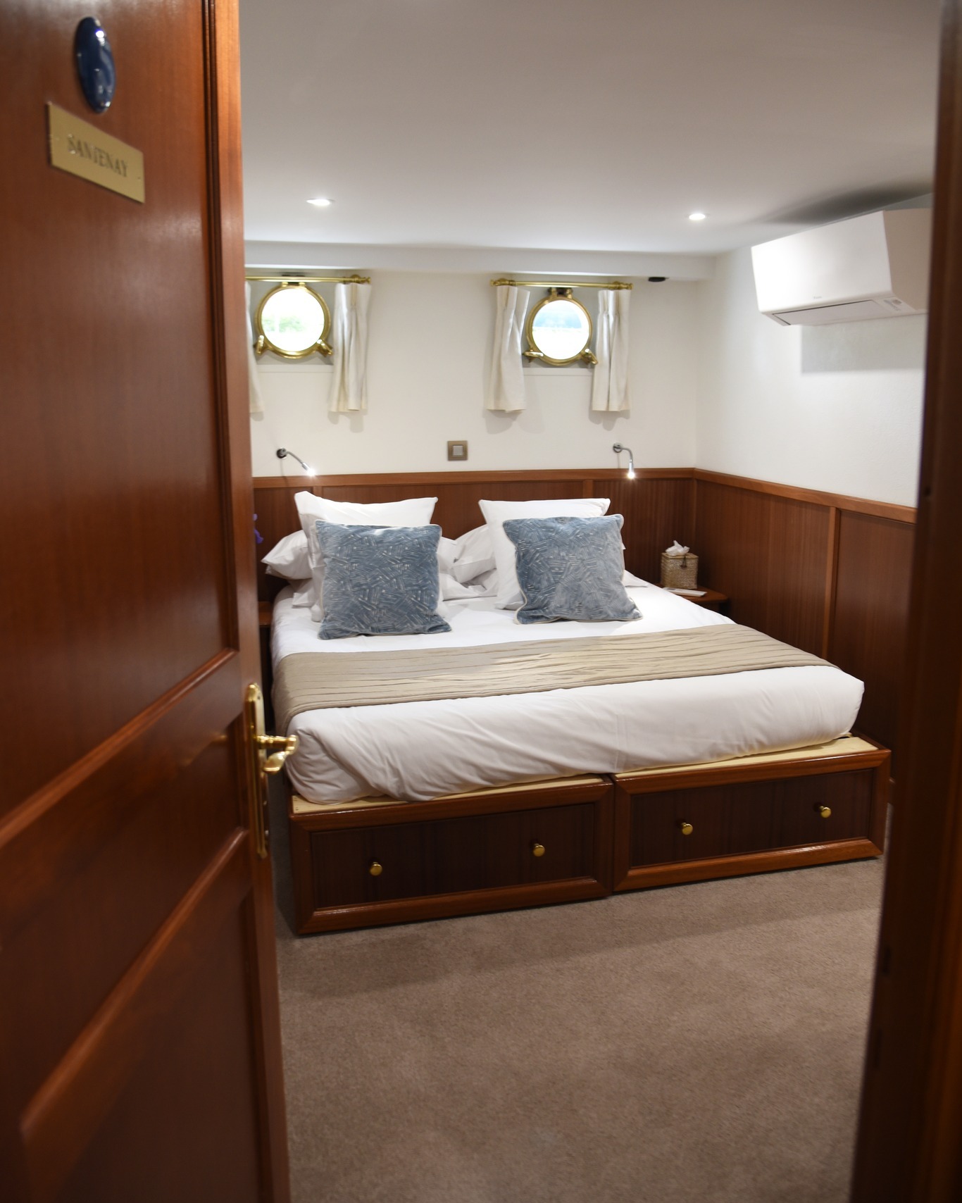 A cozy bedroom with a double bed, blue pillows, wooden drawers, and two porthole windows evokes the charm of a luxury barge cruise.