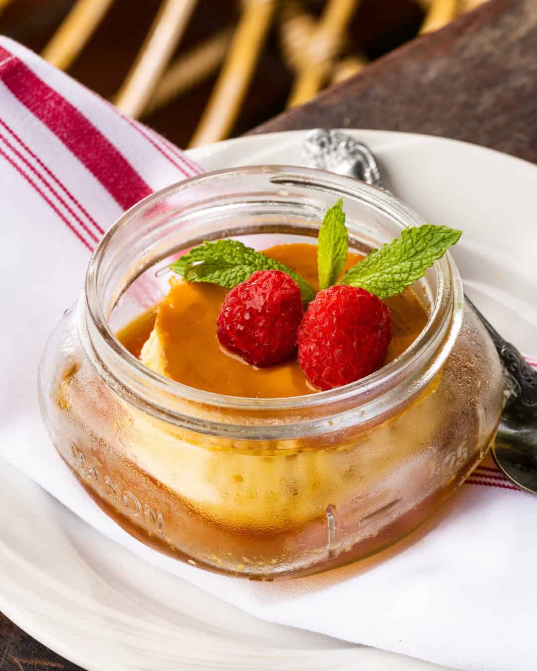 A jar of caramel flan topped with two raspberries and mint, on a plate with a red-striped napkin—a must-try treat for those who love exploring foodie destinations.