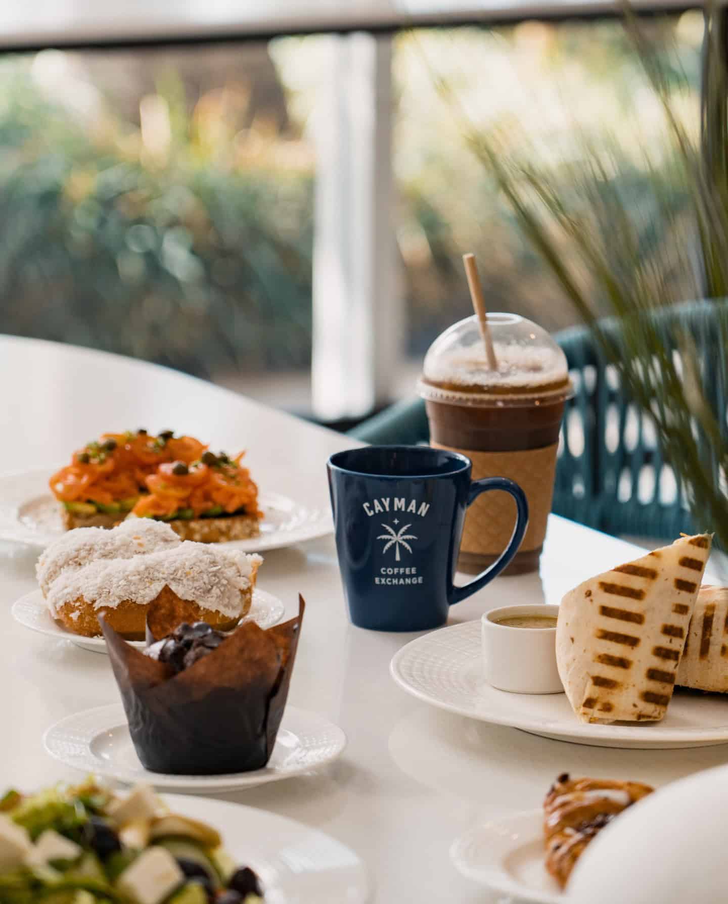 Enjoy a sunlit breakfast table set with sandwiches, pastries, a blue coffee mug, and an iced drink&mdash;perfect for starting your day in true Grand Cayman luxury travel style.