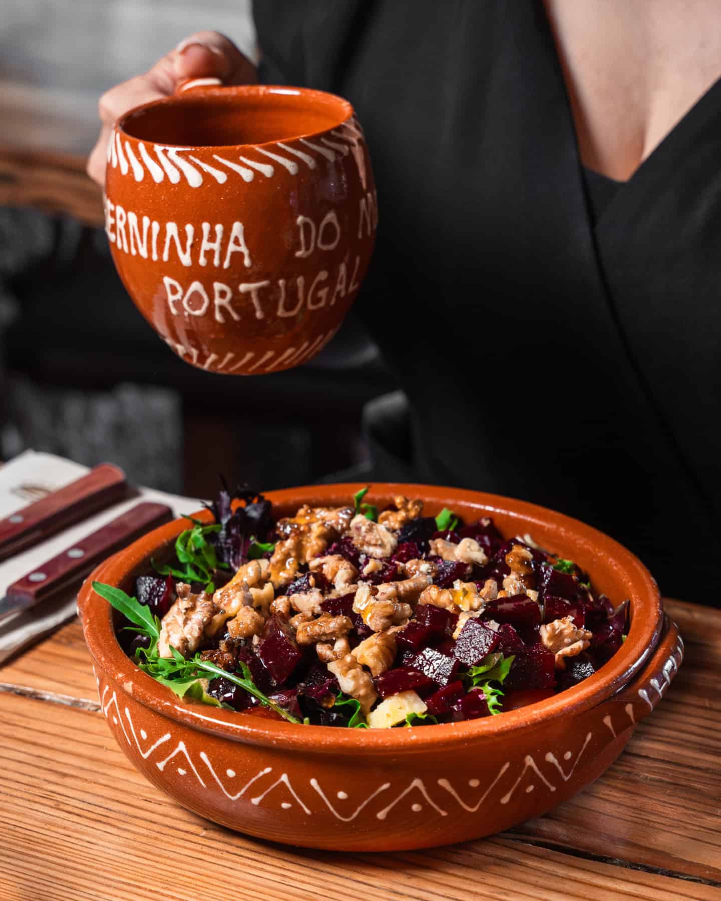 A perfect scene for Foodie Destinations: person holding a pottery mug alongside a matching bowl filled with greens, beetroot, walnuts, and cheese—an artful blend of flavor and form.