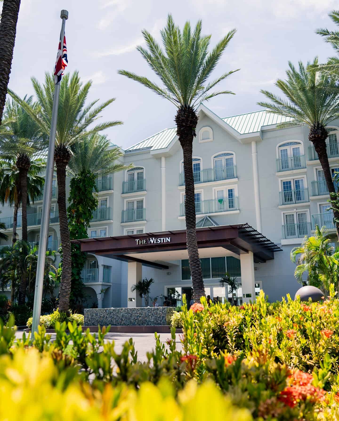 Front view of The Westin hotel with palm trees, a flagpole, and lush greenery in the foreground, offering an inviting glimpse into Grand Cayman luxury travel.