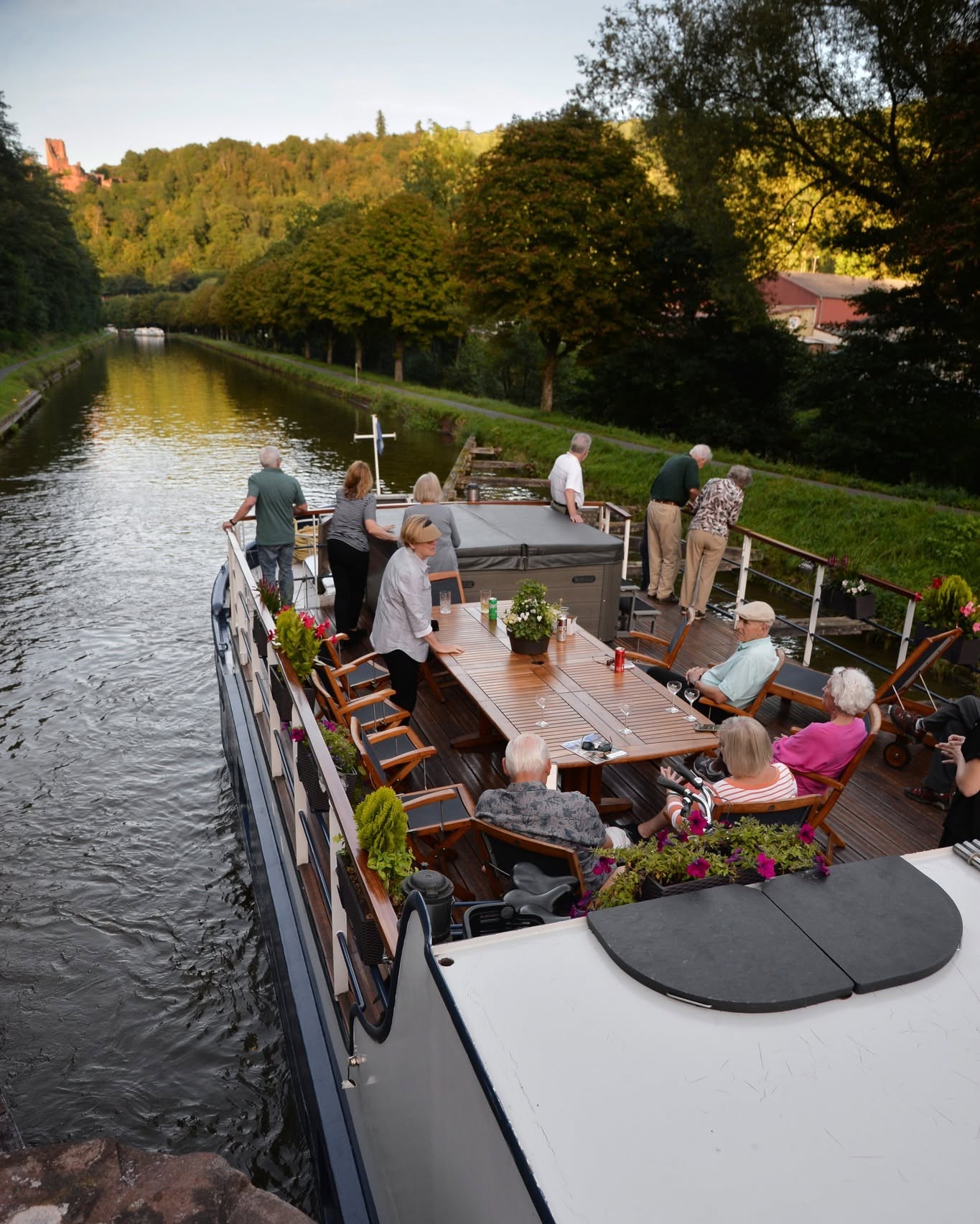 A group of people relax and socialize on the deck of a luxury barge cruise, drifting along a scenic, tree-lined river.