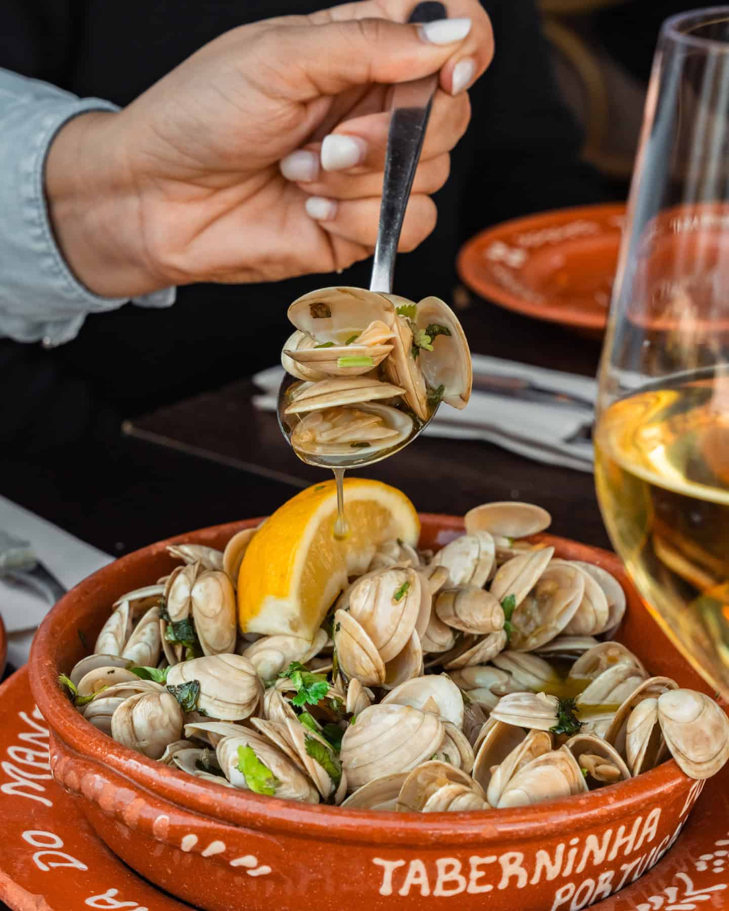 A hand holds a spoonful of clams over a clay bowl of clams with lemon and herbs, next to a glass of white wine—perfect for those seeking top foodie destinations.