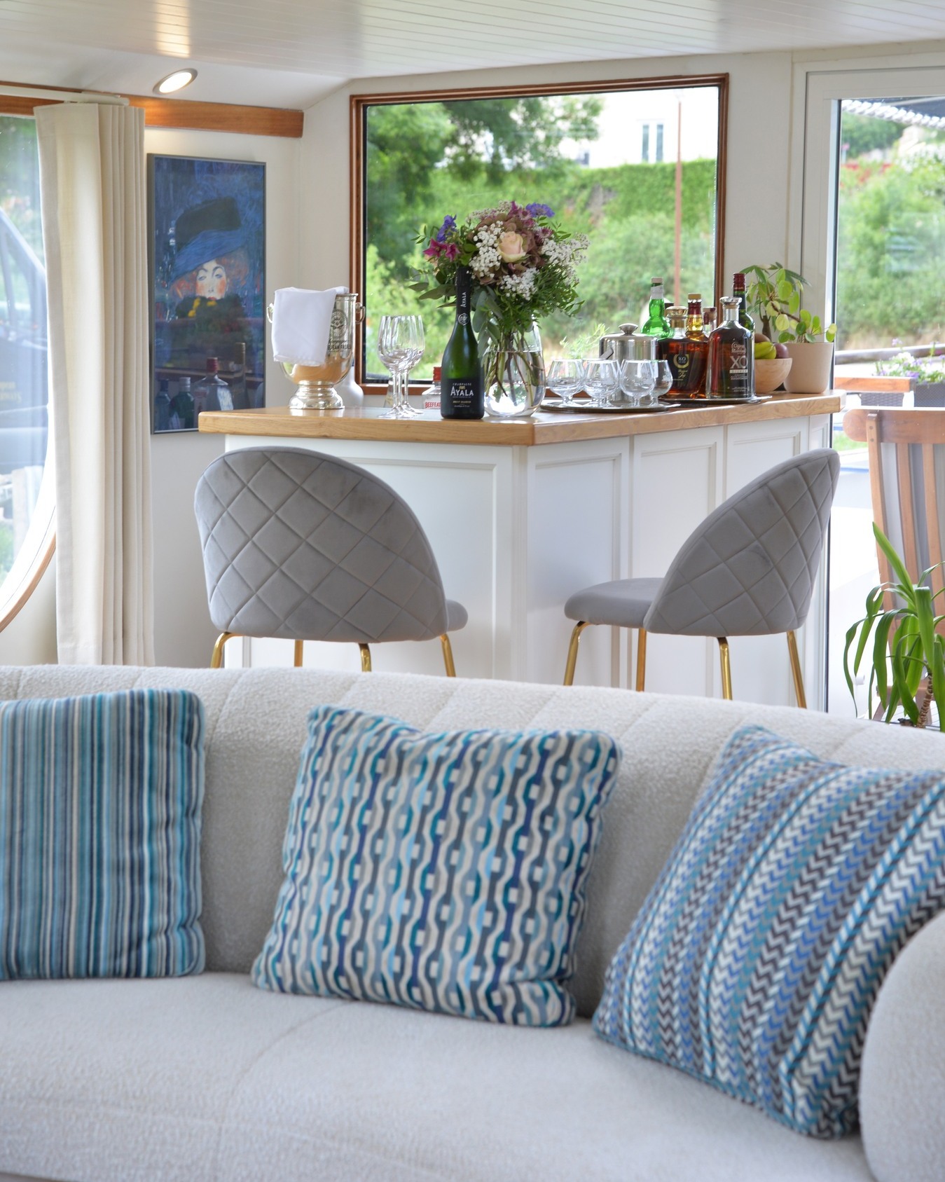 Modern living space with a bar, two gray chairs, and a white couch with patterned blue pillows—evoking the relaxed elegance of a luxury barge cruise.