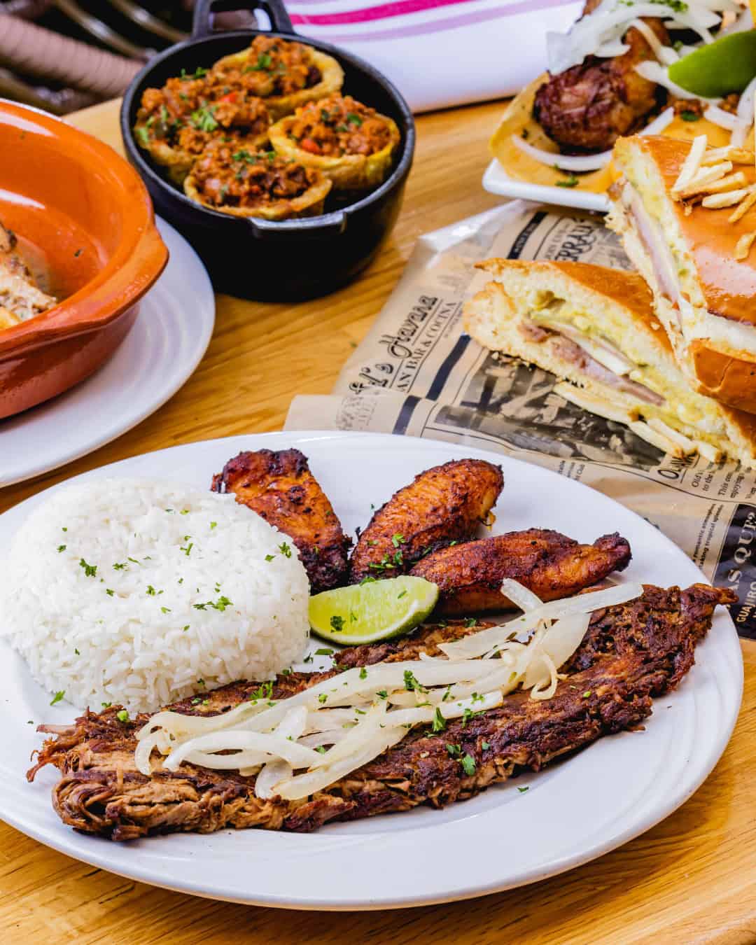 A delicious plate of white rice, grilled beef with onions, fried plantains, and lime—perfect for exploring new foodie destinations. Sandwiches and appetizers tempt in the background.