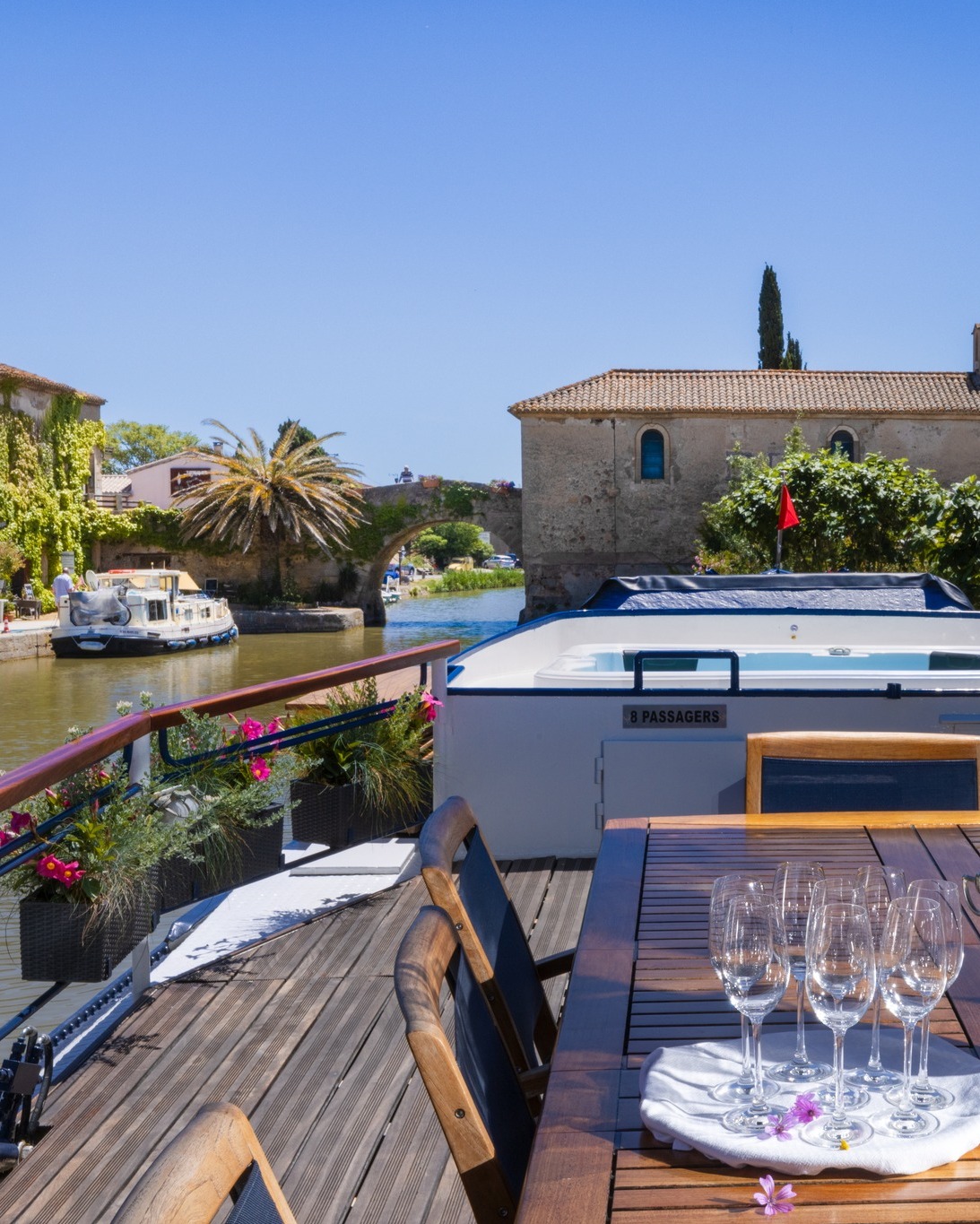Wood deck with table, empty wine glasses, flowers, canal view, boat, and buildings under blue sky—perfect for enjoying a luxury barge cruise.