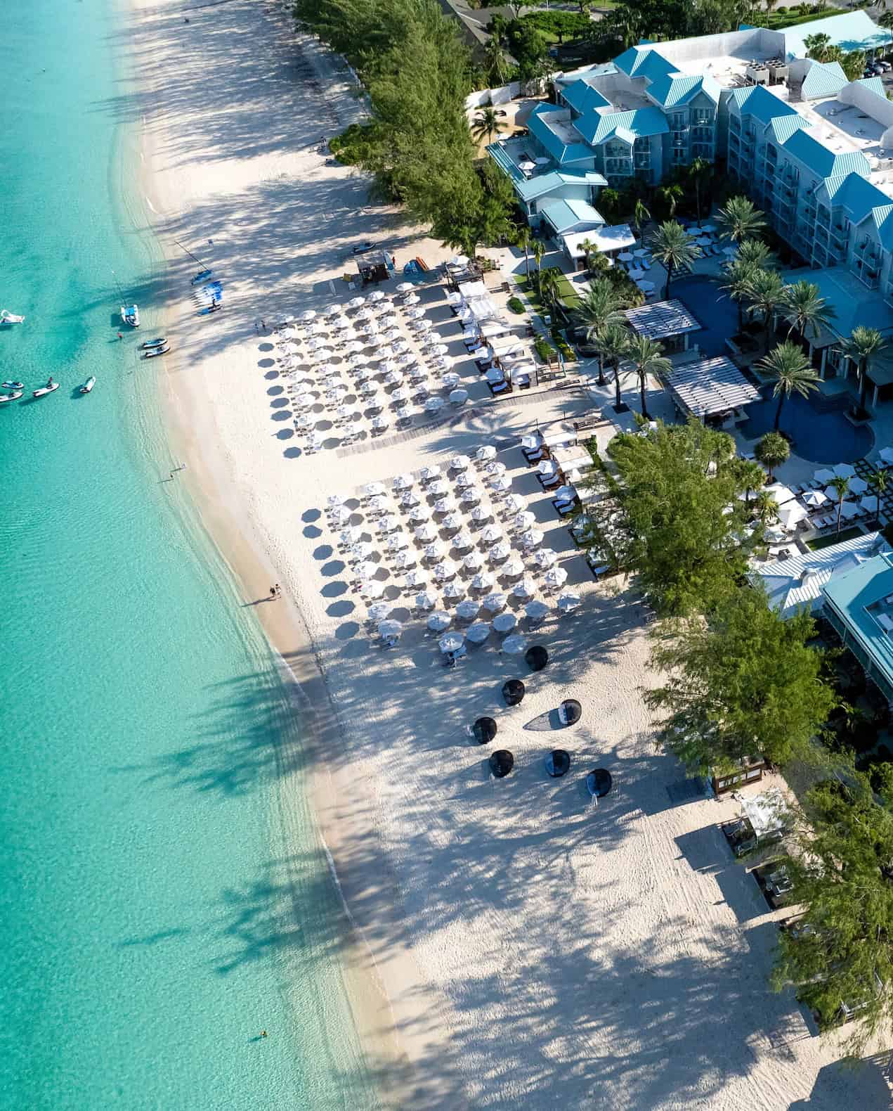 Aerial view of a tropical beach resort with umbrellas and lounge chairs on white sand by turquoise water, perfect for an unforgettable Grand Cayman Luxury Travel experience.
