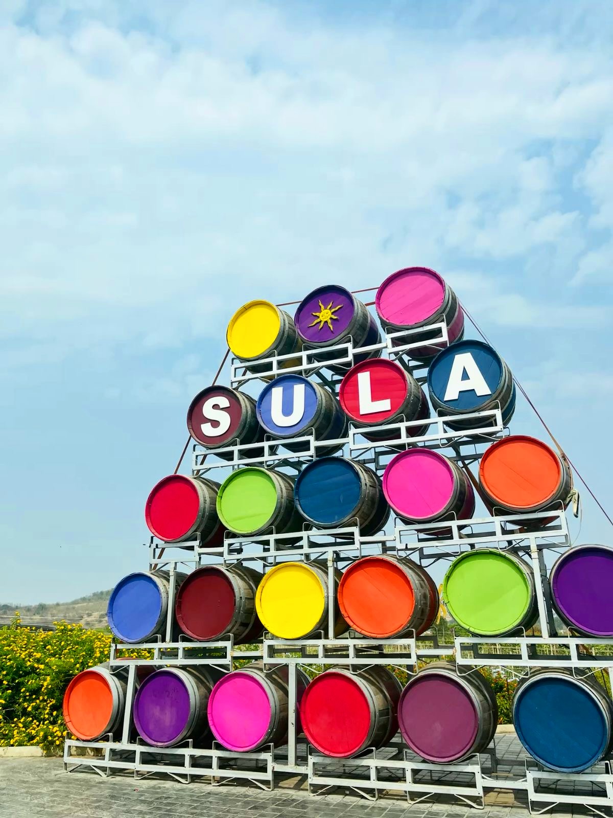Colorful barrels stacked in a pyramid shape with "SULA" spelled out near the top, outdoors under a blue sky&mdash;a vibrant symbol of wine tourism.