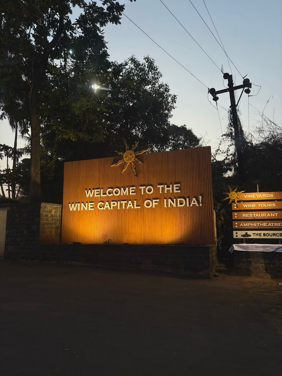 A large illuminated sign reads "Welcome to the Wine Capital of India!" beside direction boards at dusk, inviting visitors to explore the region's thriving wine tourism.