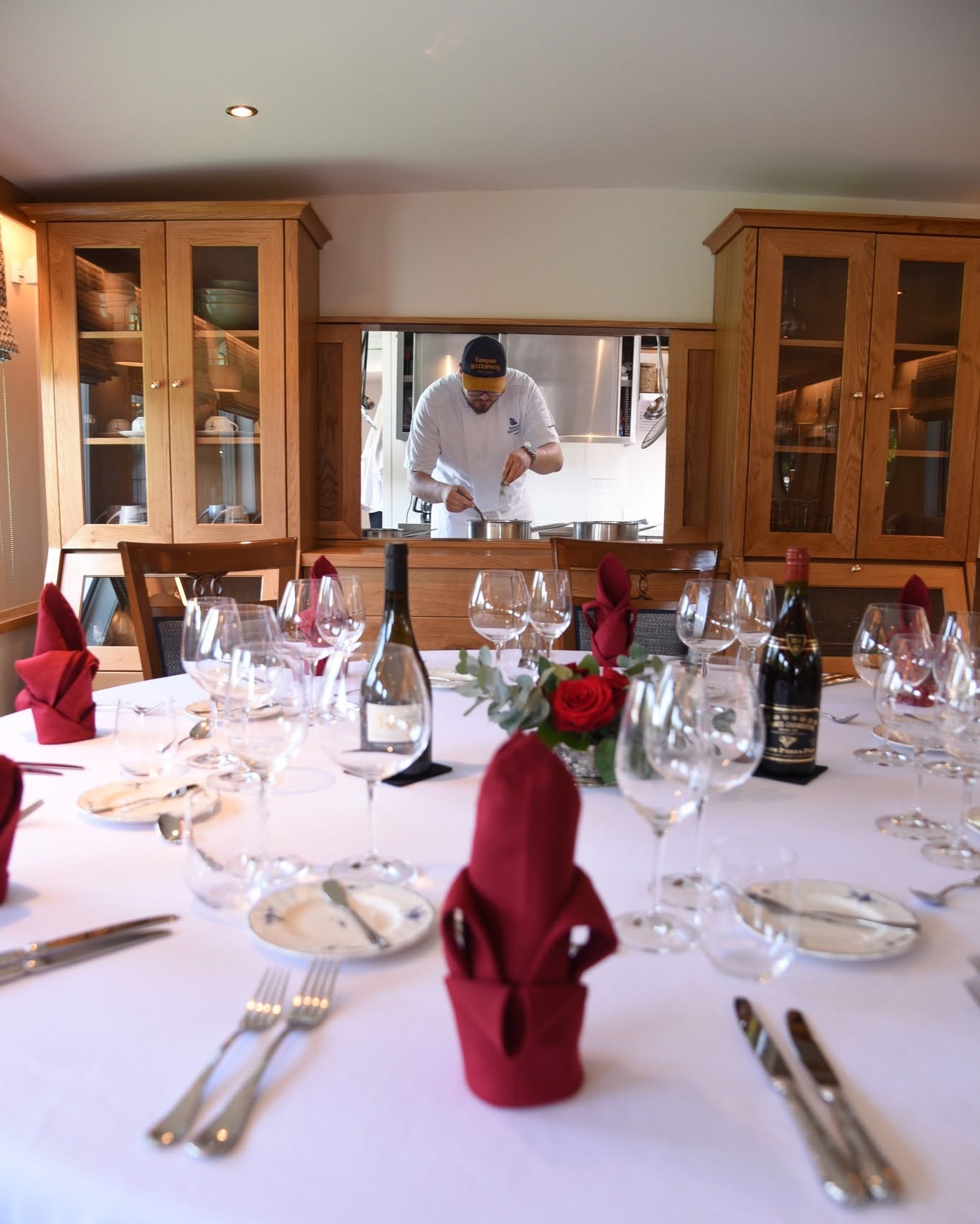 Chef preparing food in the kitchen, seen through a window behind an elegantly set dining table, evokes the refined ambiance of a luxury barge cruise.