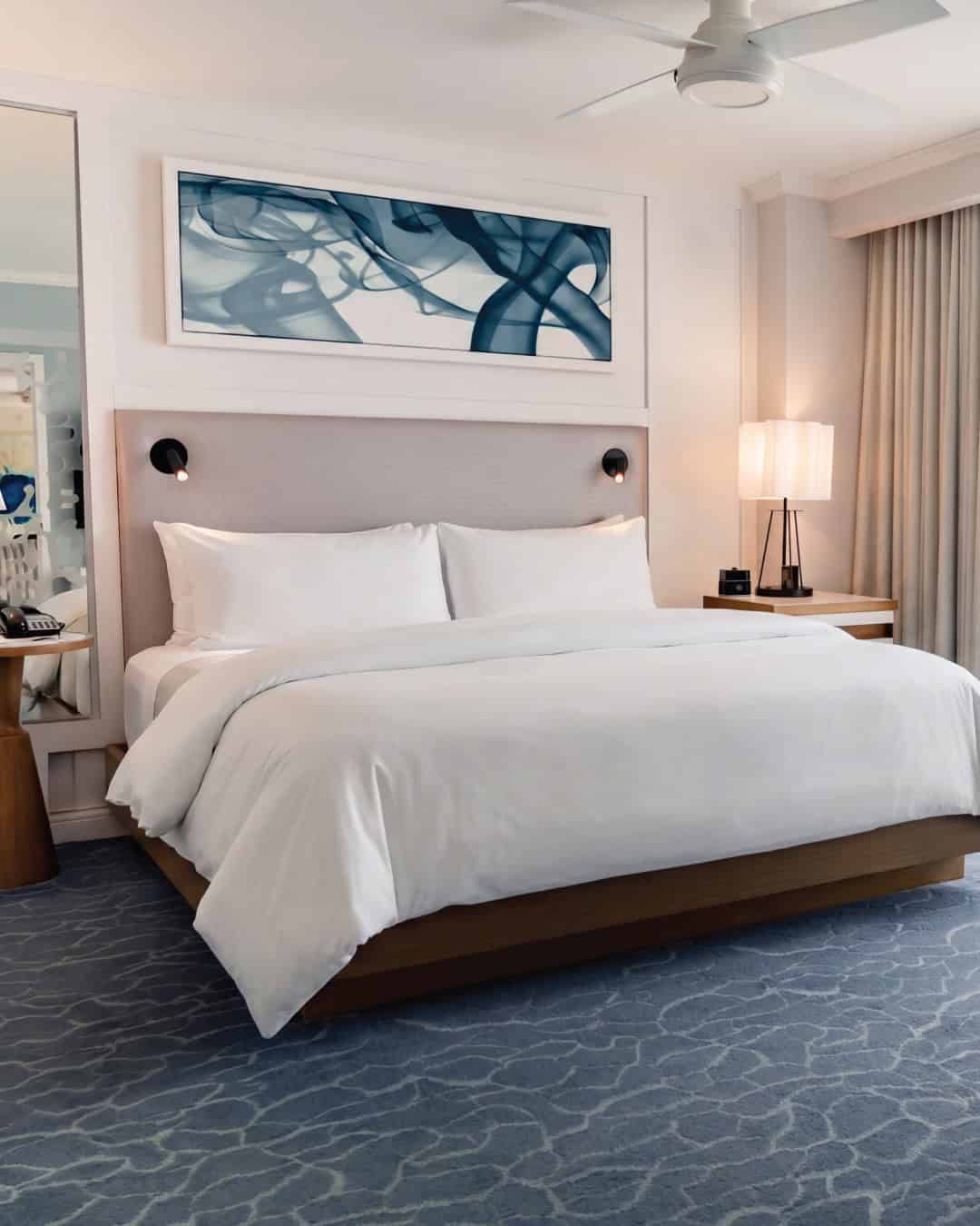 A modern hotel room designed for Grand Cayman luxury travel, featuring a large bed, abstract blue art above the headboard, a ceiling fan, and an elegant blue patterned carpet.