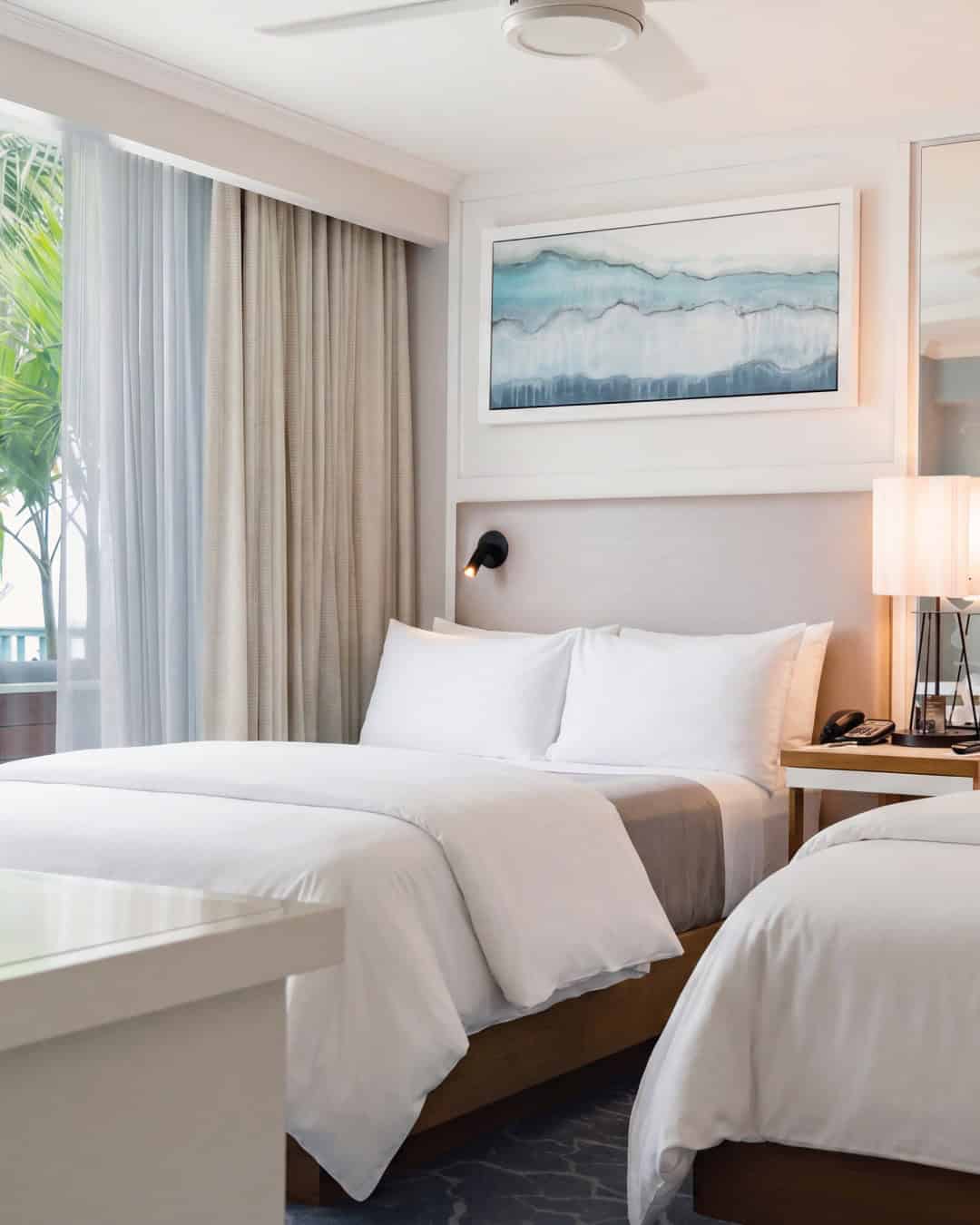 Modern hotel room with two white beds, abstract wall art, lamp, phone, and large window with curtains&mdash;perfect for Grand Cayman luxury travel.