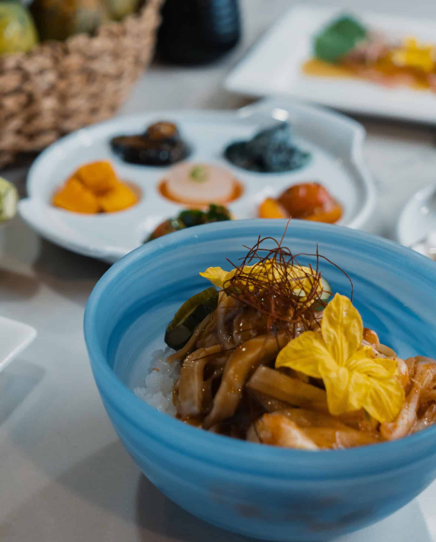 A blue bowl with rice, vegetables, and edible flowers offers a taste of Grand Cayman luxury travel, with assorted dishes blurred in the background.