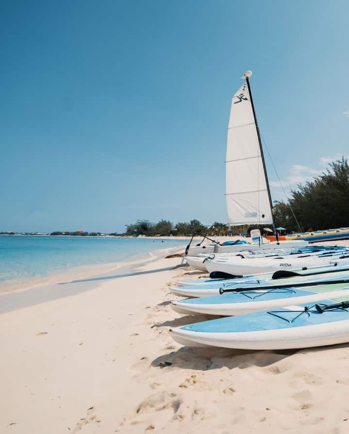 Paddleboards and a small sailboat lined up on a sandy beach by clear blue water under a sunny sky set the perfect scene for Grand Cayman luxury travel.