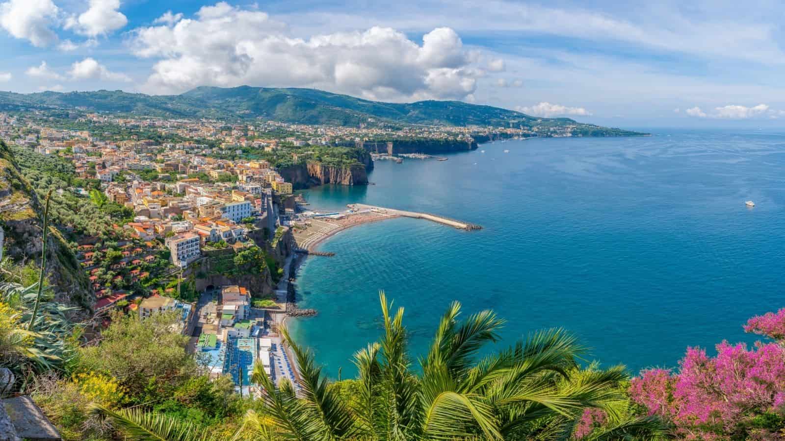 Coastal town with colorful buildings, a marina, lush greenery, and mountains by a bright blue sea under clouds—one of the most scenic coastal destinations and wonderfully easy to explore on foot with its car-free travel atmosphere.