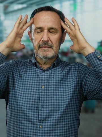 A man in a plaid shirt holds his head with both hands, appearing stressed at an airport terminal.