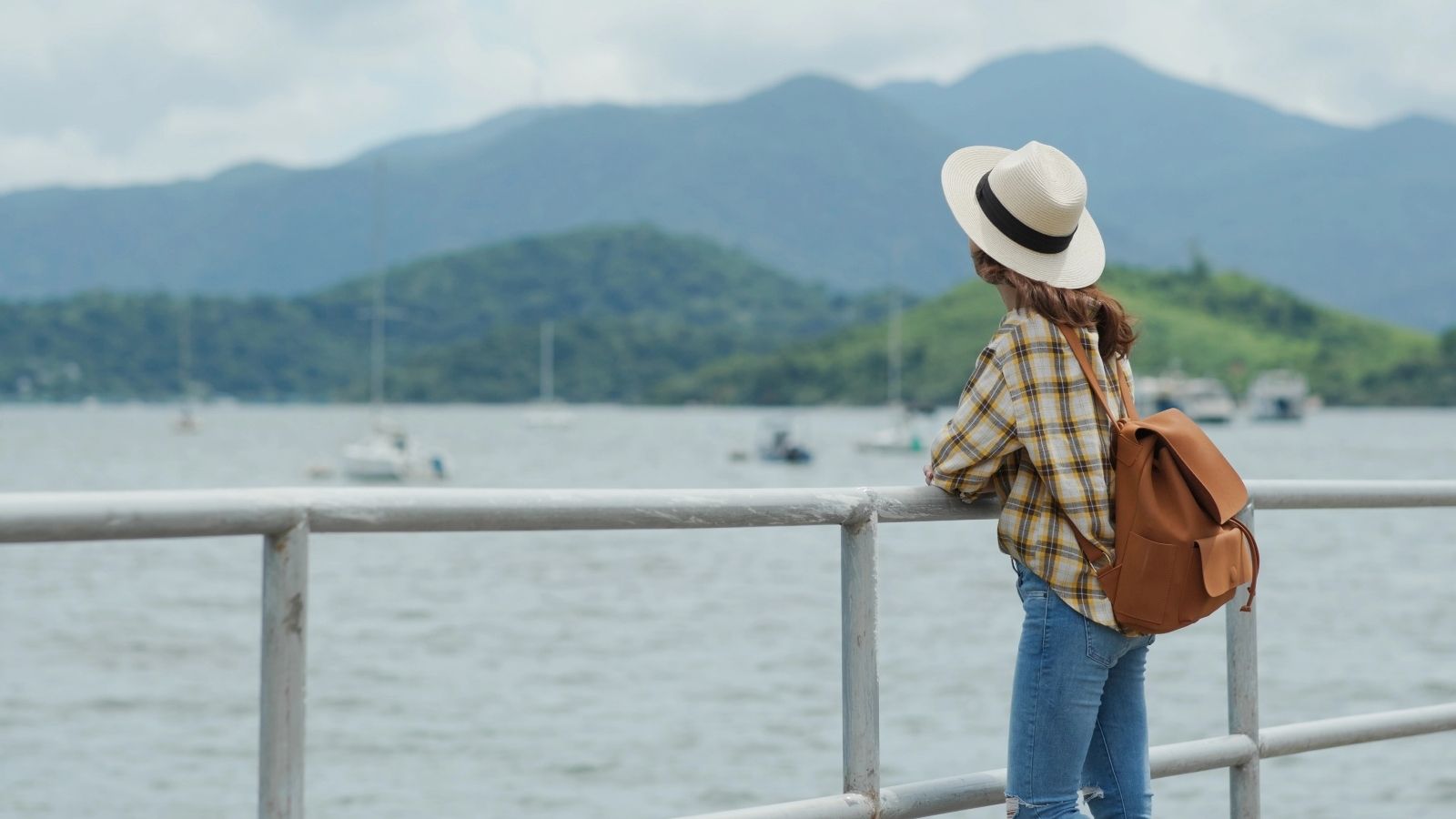 Woman in hat and backpack stands by railing, looking at mountains and water with boats in the distance.