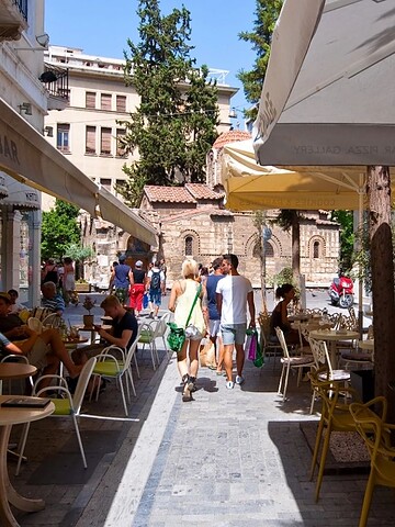 People walk past outdoor cafes on a sunny street with tables, chairs, and shade umbrellas.