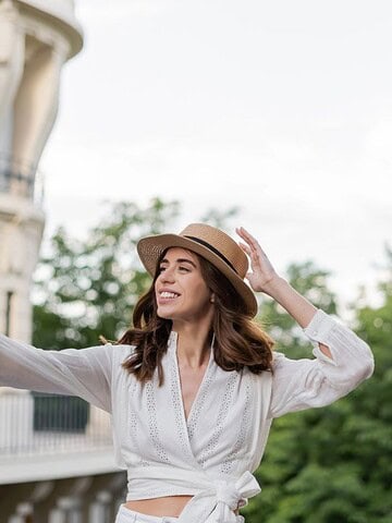 Woman in a white blouse and hat takes a selfie on a balcony with the Eiffel Tower in the background.