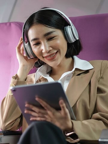 Woman wearing headphones smiles while using a tablet, seated on a plane with purple seats.