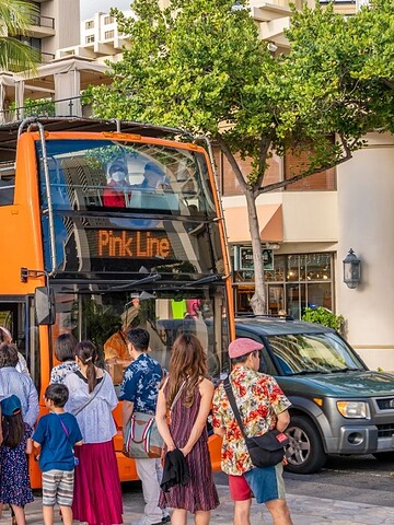 People in summer clothes line up to board a bright orange Waikiki Trolley bus in a city setting.