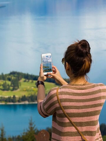 Woman taking a photo of a scenic lake and town, with paragliders in the sky, on a sunny day.