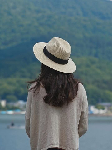 A woman in a hat stands by a lake, facing green mountains in the distance.
