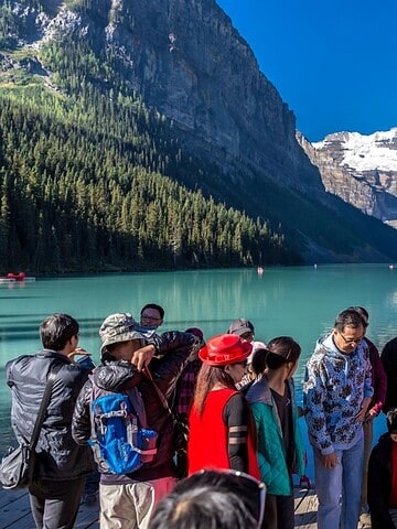 A group of tourists gather by a turquoise lake with snowy mountains and pine trees in the background.