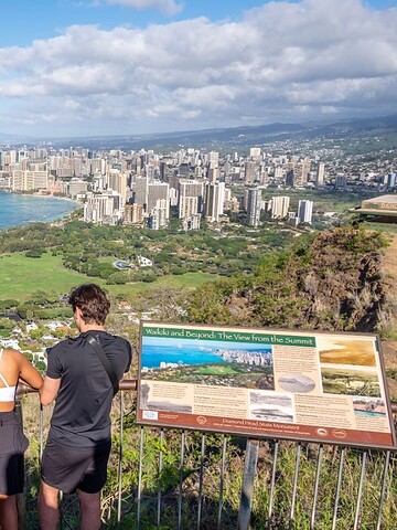 People at a scenic lookout view Honolulu skyline and the ocean from a mountain viewpoint on a sunny day.
