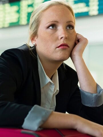 Woman in business attire looking bored, resting her head on her hand with a suitcase at an airport.