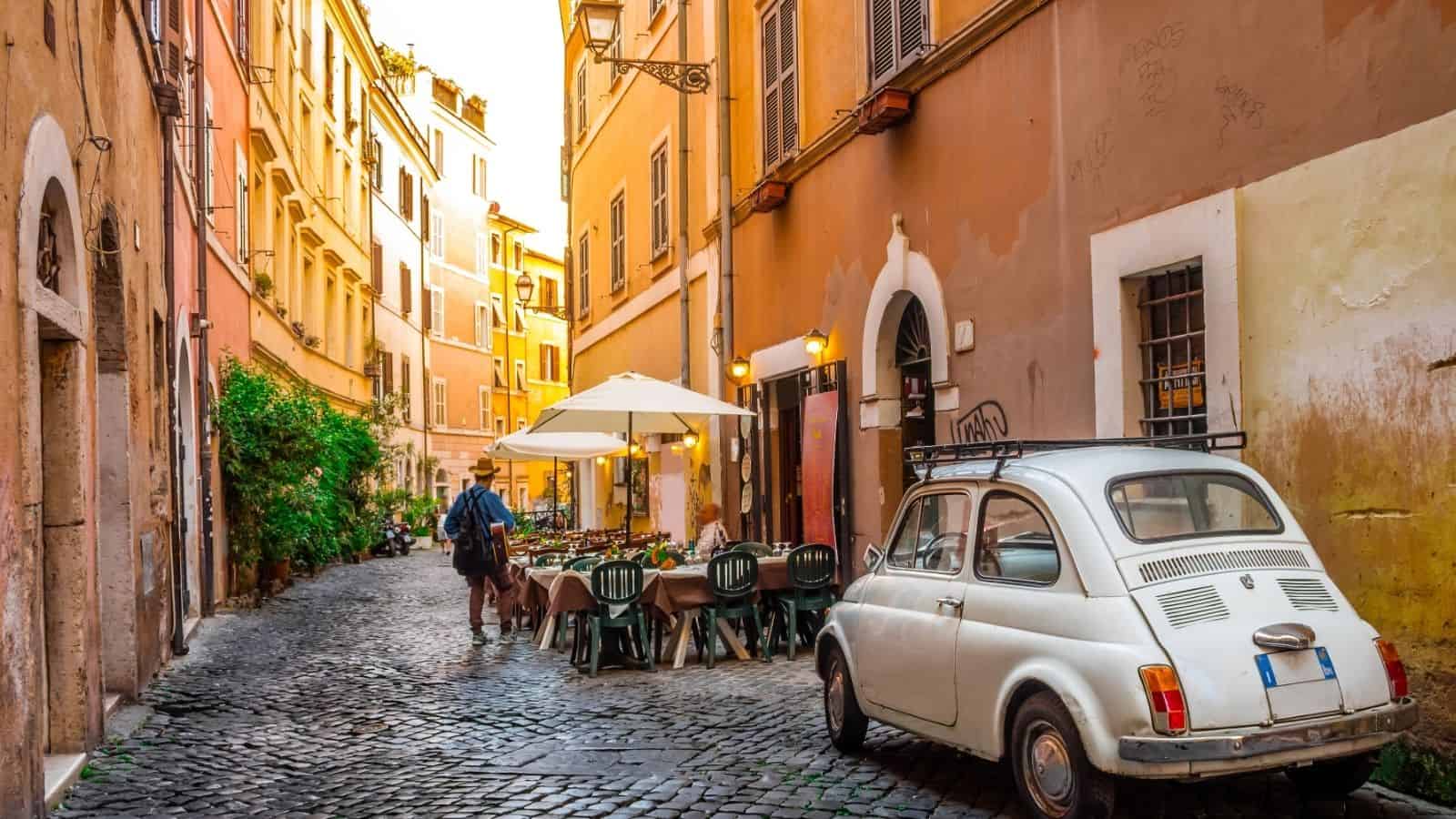 A narrow, cobblestone street with a small vintage car, outdoor caf&eacute; seating, and warm-toned buildings.