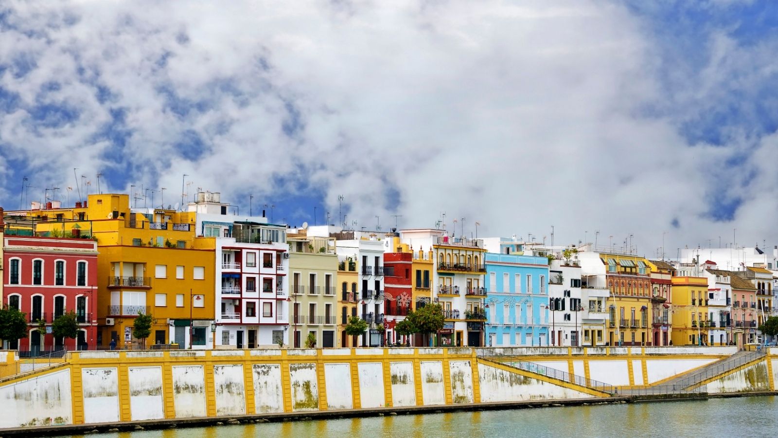 Colorful buildings line a river under a cloudy sky, with reflections visible in the water.
